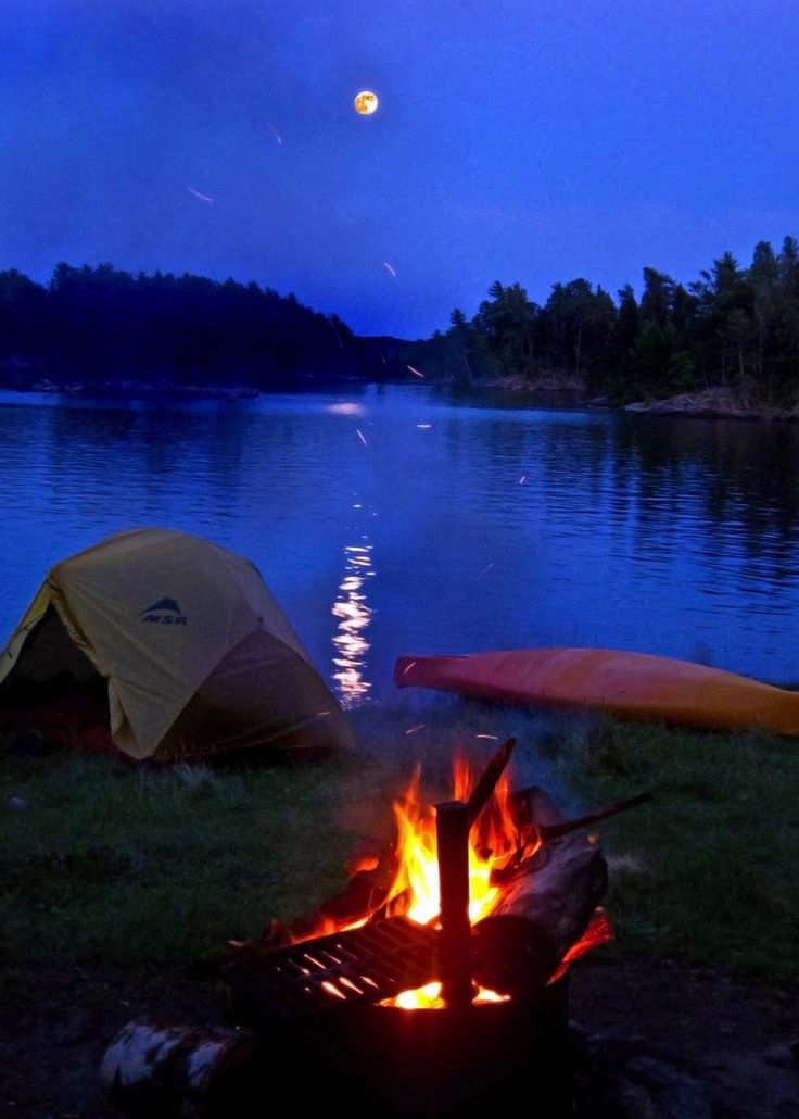 two tents set up next to a campfire on the shore of a lake at night