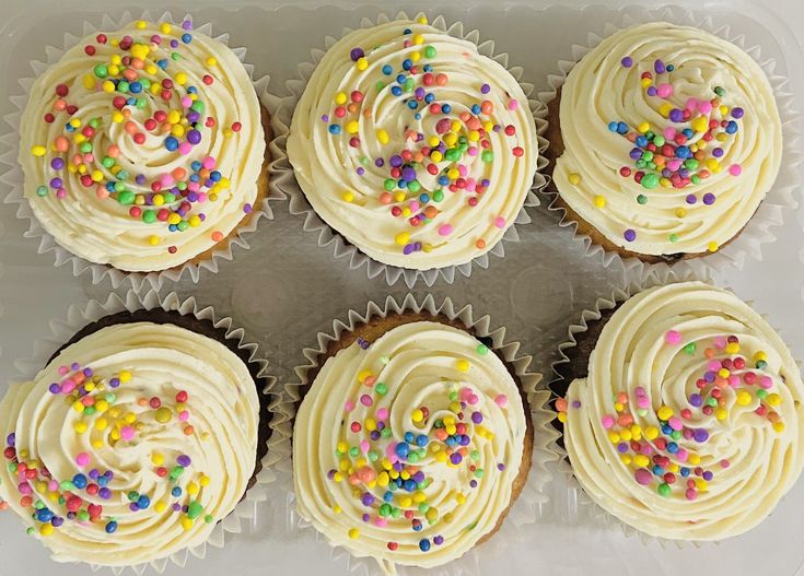 six cupcakes with white frosting and sprinkles in a plastic container