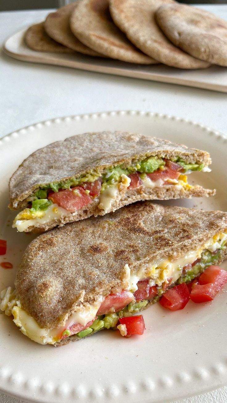 two sandwiches on a plate with tomatoes and other food items in the background, along with pita bread