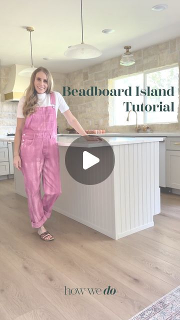 a woman in pink overalls standing next to a kitchen island with white cabinets and drawers