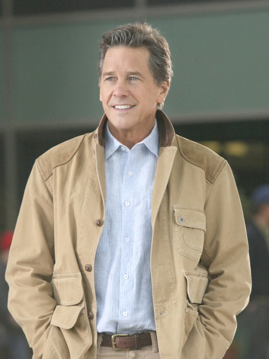 a man standing in front of a building wearing a brown jacket and tan pants with his hands in his pockets