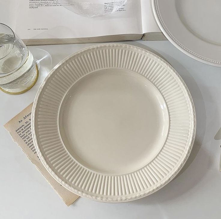 an empty white plate next to a glass and book on a table with utensils