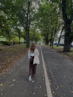 a woman walking down the middle of a tree lined road in an open park area