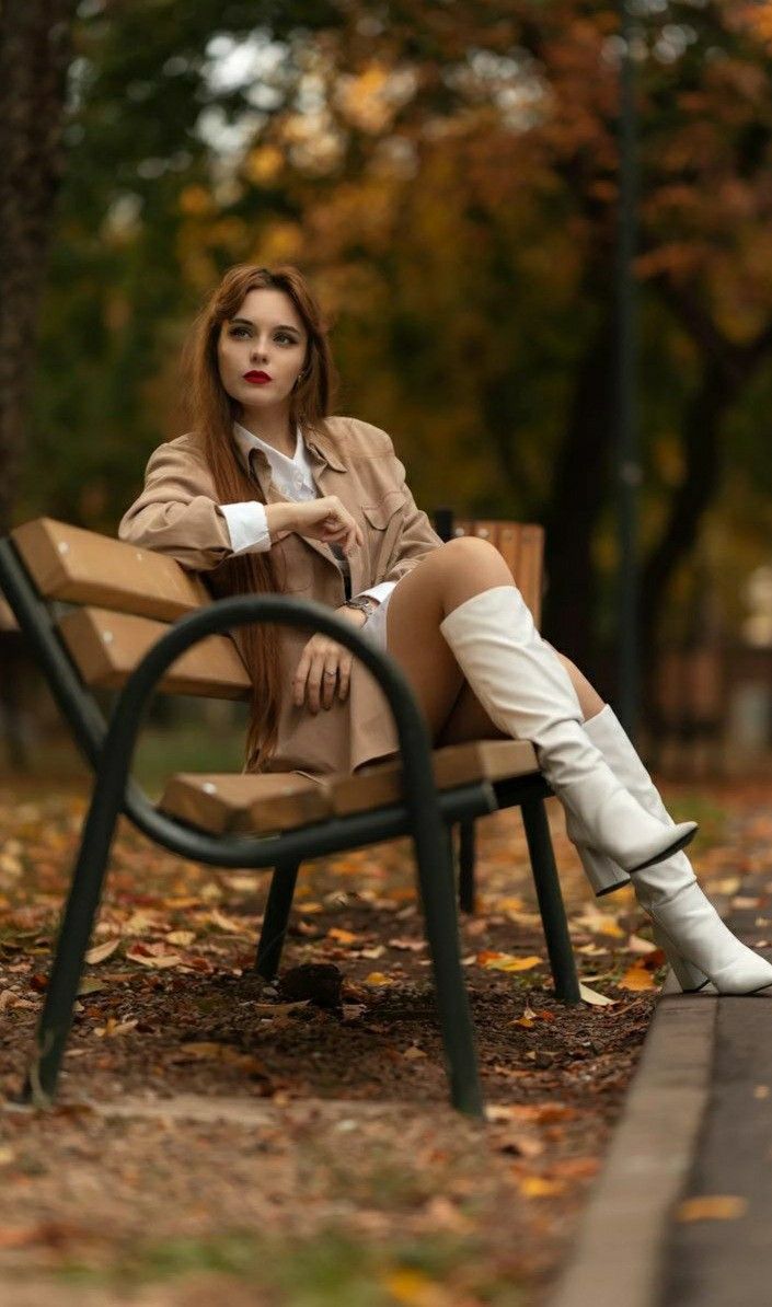 a woman is sitting on a bench in the park with her legs crossed and wearing white boots