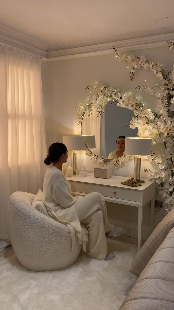 a woman sitting on a chair in front of a mirror and vanity with flowers around it