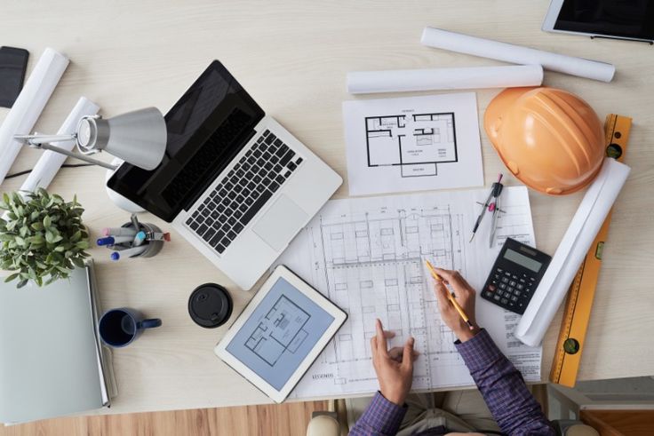 a person sitting at a desk working on architectural drawings and laptops next to other items