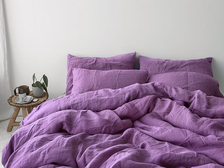 an unmade bed with purple linens in a white room next to a window