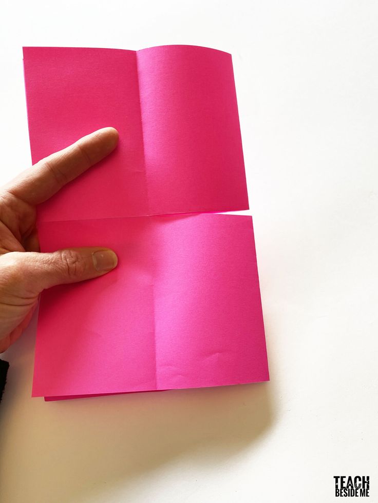 a hand is pointing at the corner of a piece of pink paper that has been folded in half