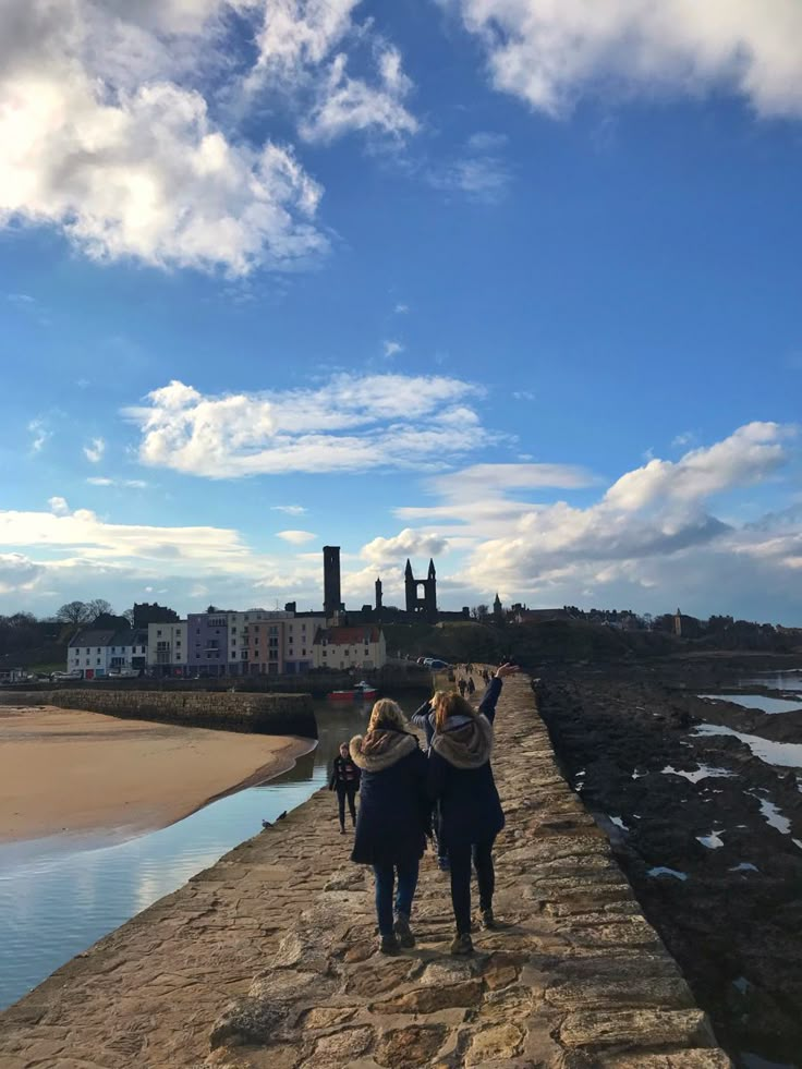 two people are walking along a stone wall near the water and buildings in the background