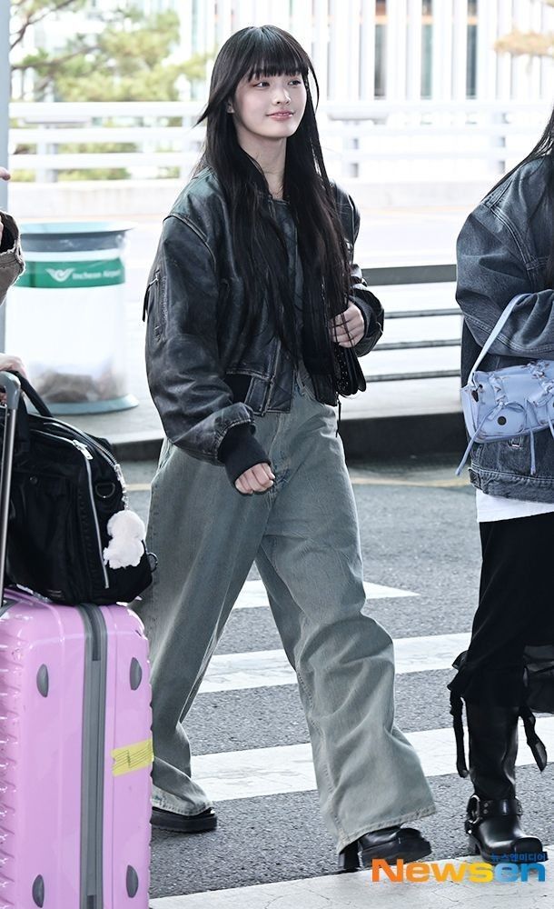 a young woman walking across an airport with her pink suitcase and black jacket over her shoulders