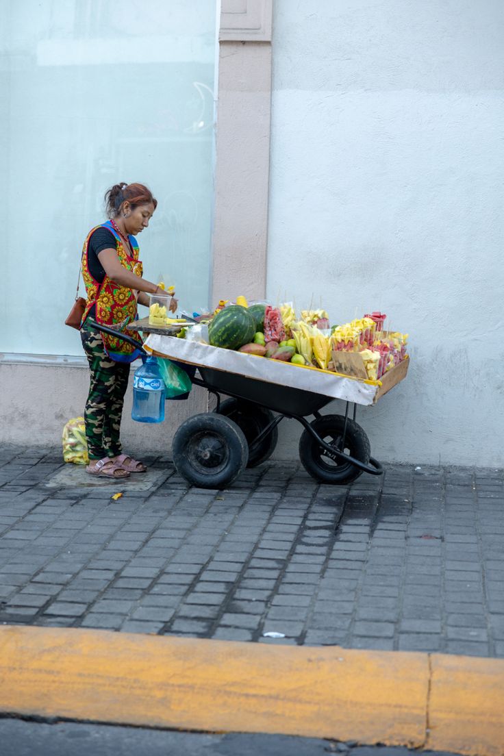 a woman standing next to a wheelbarrow filled with fruits and vegetables