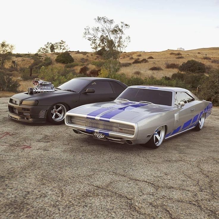 two muscle cars parked next to each other on a dirt road in the middle of nowhere