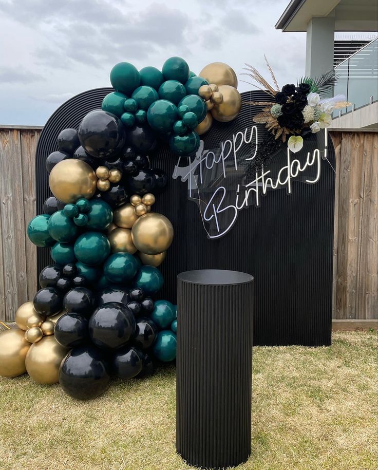 a black and gold balloon arch with happy birthday written on it in front of a wooden fence