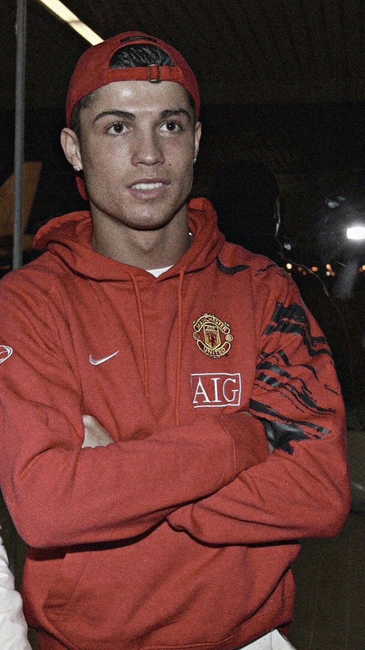 a young man in a red sweatshirt and cap standing with his arms crossed looking at the camera