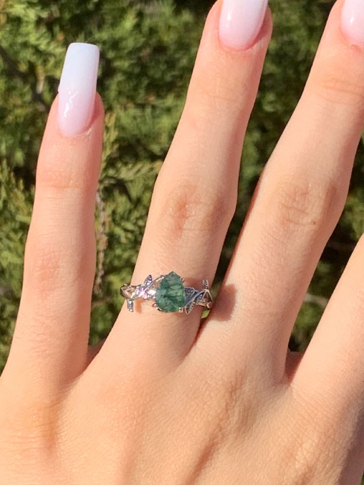 a woman's hand with a ring on top of her finger and some green plants in the background