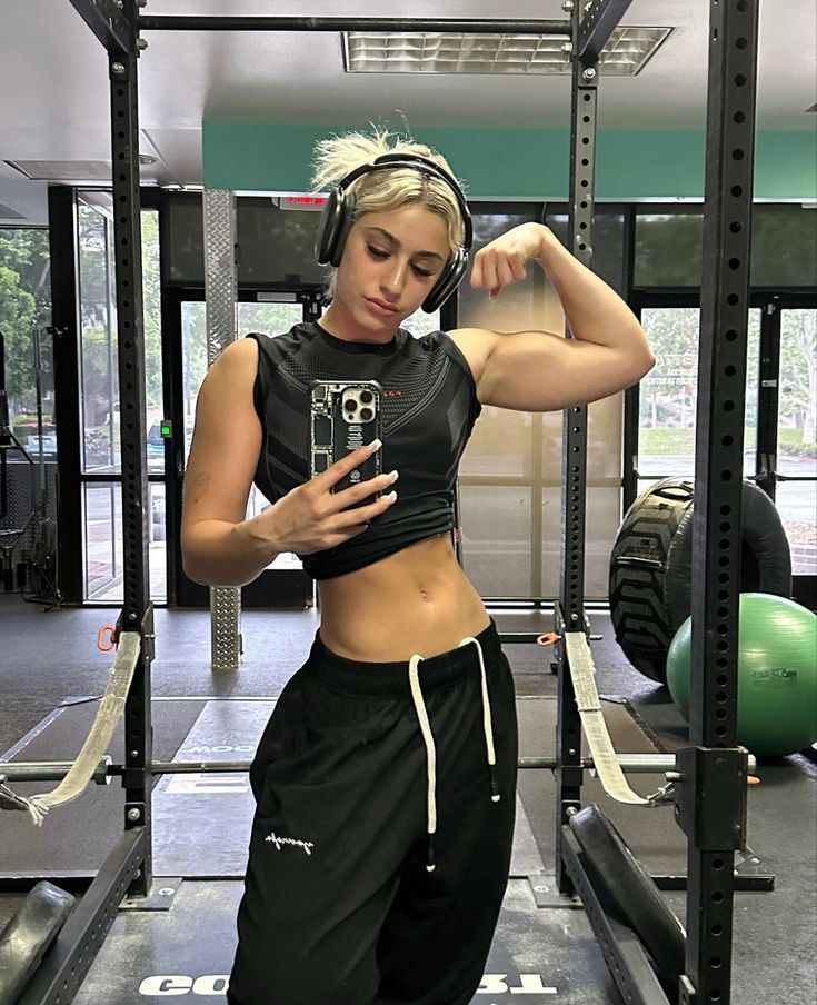 a woman standing in the middle of a gym with headphones on and looking at her cell phone