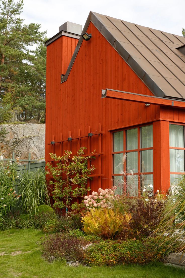 a red house with a metal roof in the middle of some grass and flowers around it