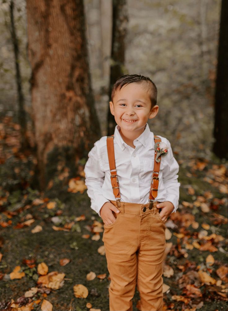 a young boy wearing suspenders and a white shirt smiles at the camera while standing in front of some trees