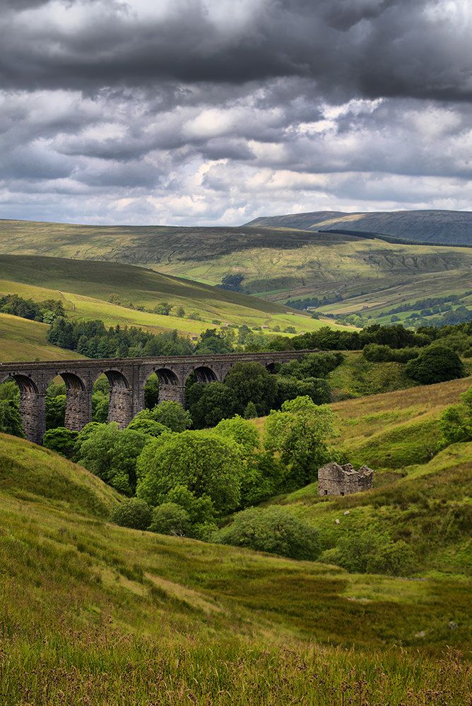 an old train traveling over a bridge in the middle of a lush green valley under a cloudy sky