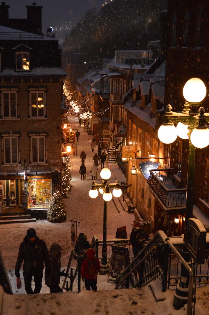 people walking down a snowy street at night with lights on and buildings in the background