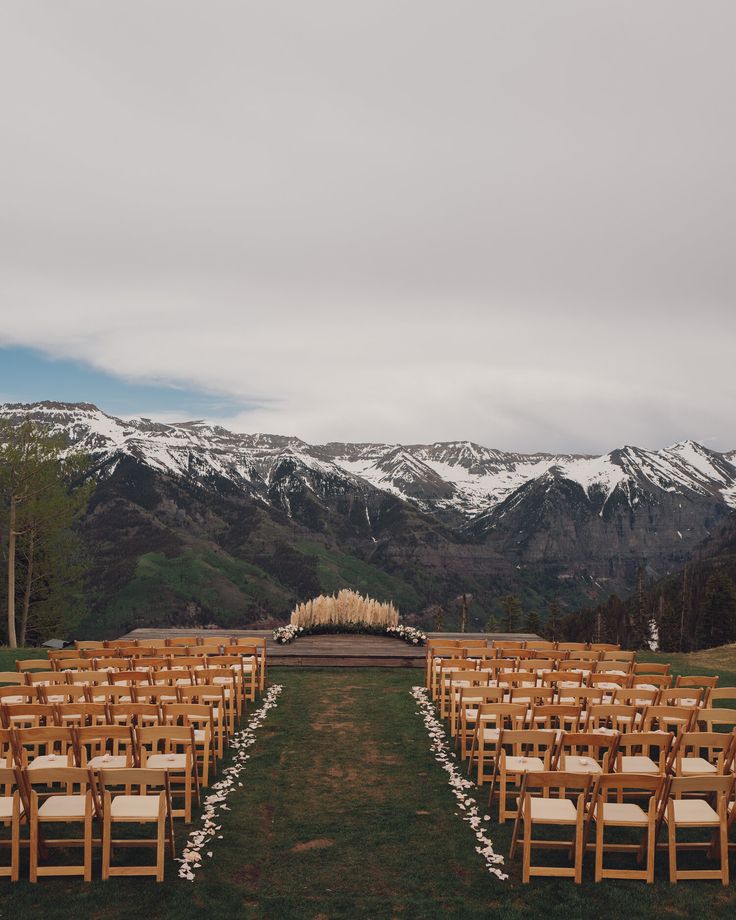 an outdoor ceremony setup with wooden chairs and white flowers on the aisle, in front of snow - capped mountains