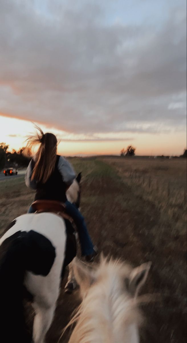 two people riding horses on a dirt road at sunset or dawn with the sun setting in the distance