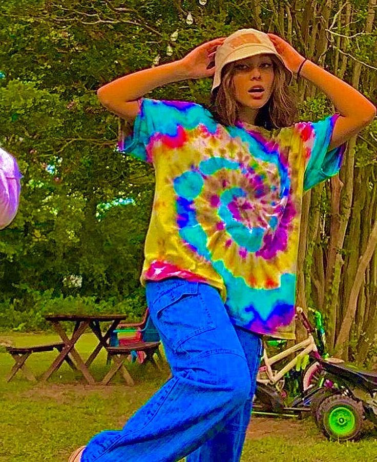 a young woman in a tie - dyed shirt and blue jeans is dancing with her hands on her head