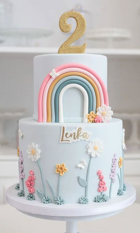 a two tiered cake with flowers and a rainbow on the top that says lenka