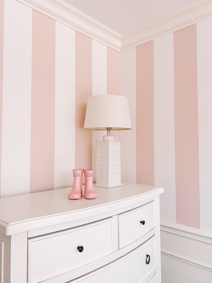 a white dresser with two pink vases on top and a lamp next to it