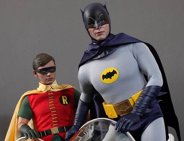 batman and robin wayne action figures on display in a museum setting with grey background, two men dressed as superheros