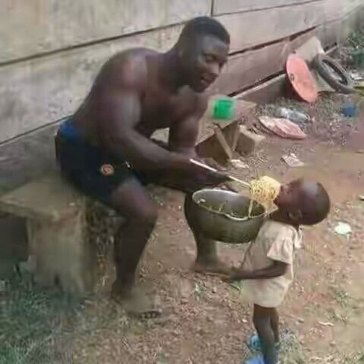 a man is feeding a small child some food from a bucket on the ground near a building