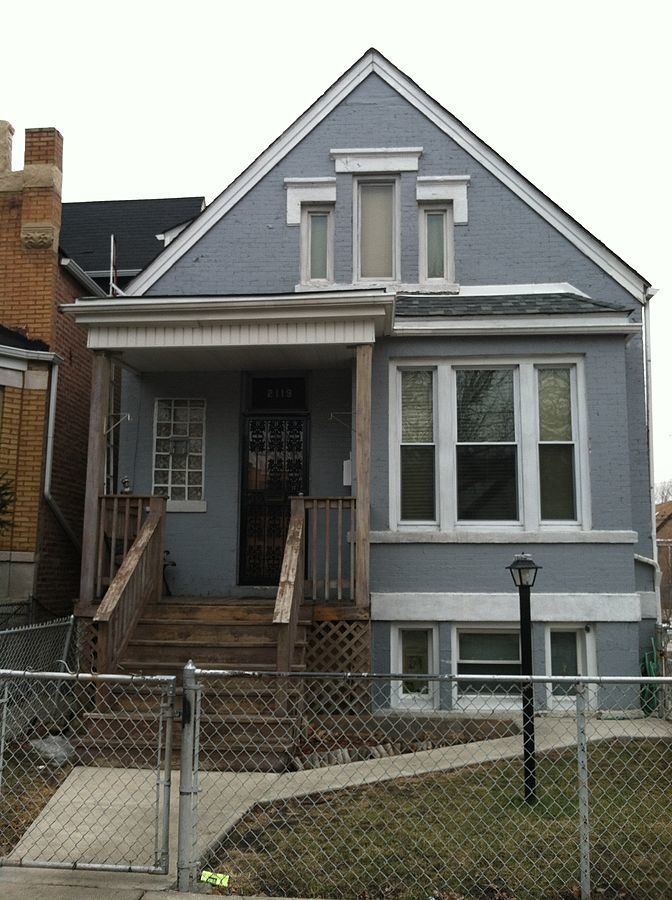 a blue house with white trim and two story houses in the background, behind a chain link fence