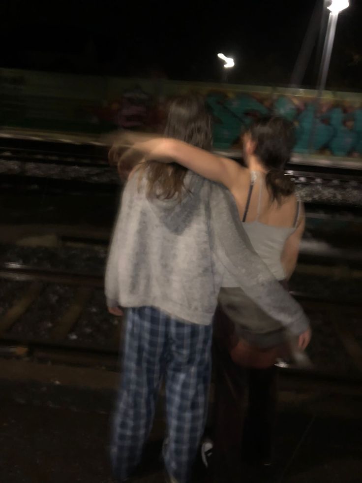 two people standing next to each other in front of a train track at night time