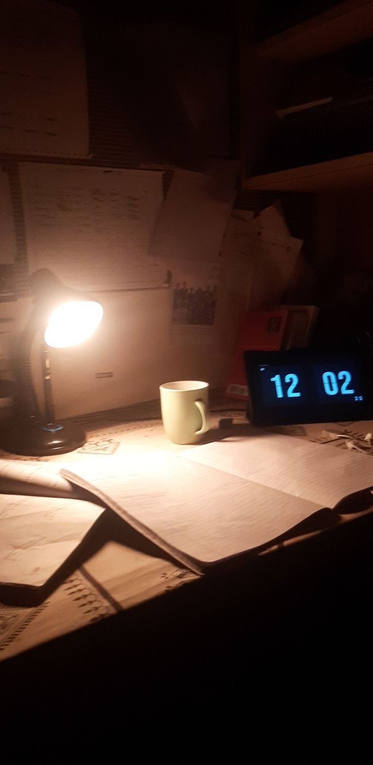 an alarm clock sitting on top of a desk next to a cup and lamp in the dark