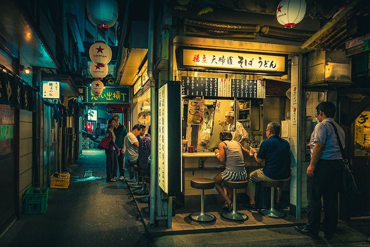 some people are sitting at a small bar in the middle of an alleyway with lanterns hanging from the ceiling