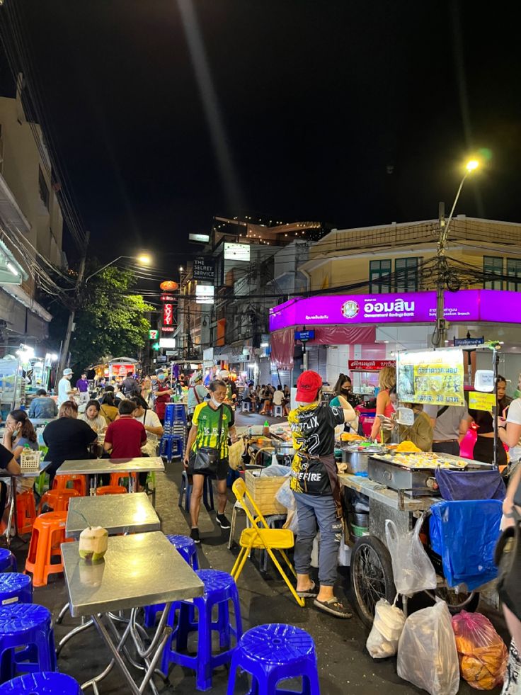 a group of people standing around tables and chairs on a city street at night with buildings in the background