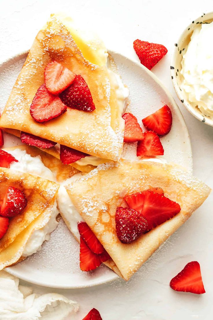 strawberry crepes with whipped cream and sliced strawberries on a plate next to a bowl of whipped cream