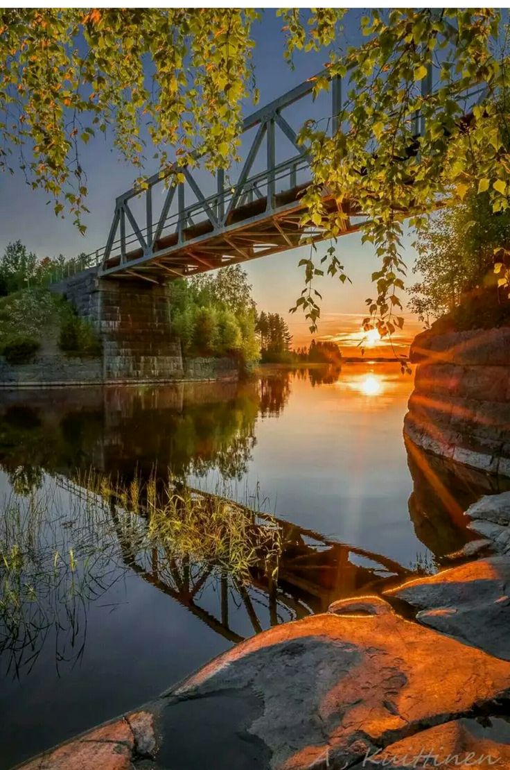 the sun is setting behind a bridge over a body of water with rocks in front of it