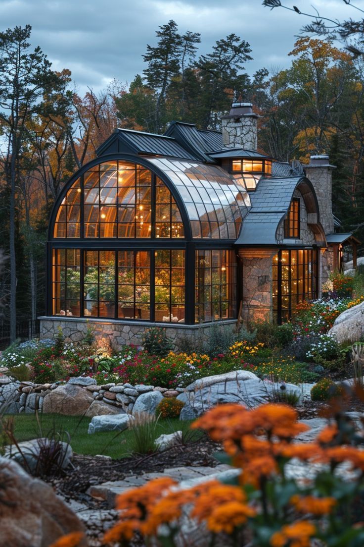 a glass house surrounded by rocks and flowers in the evening time with lights shining on it's windows