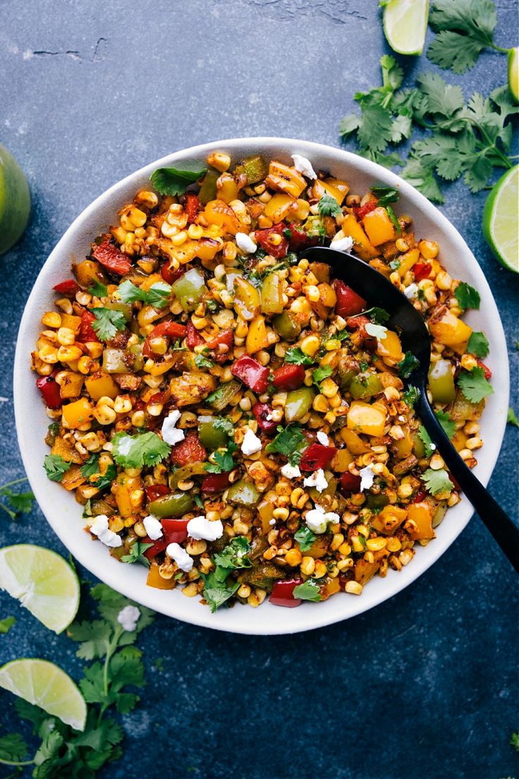 a white bowl filled with mexican corn salad next to limes and cilantro