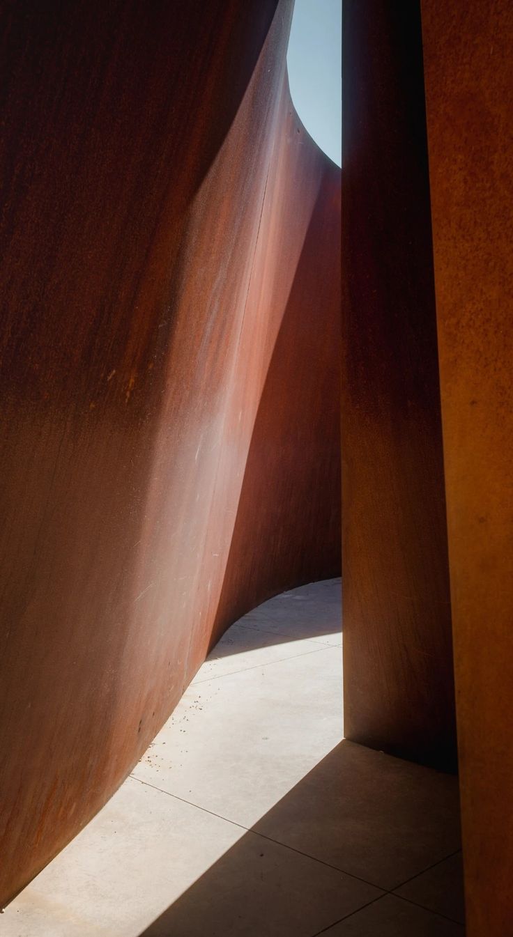 the sun shines through two tall, curved metal structures in an open space with concrete flooring