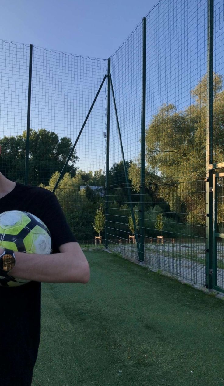 a man holding a soccer ball in front of a fenced area with grass and trees