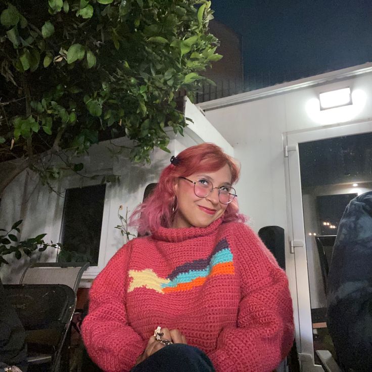 a woman with pink hair and glasses sitting on a chair in front of a house