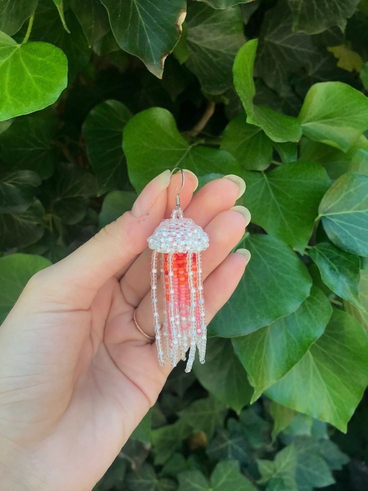 a hand holding an orange and white beaded dream catcher in front of green leaves