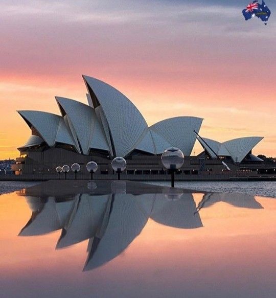 the sydney opera house is reflected in the water at sunset, with an airplane flying overhead