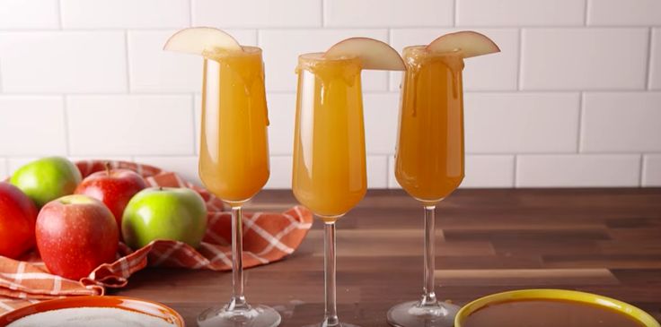 three glasses filled with orange juice on top of a wooden table next to an apple