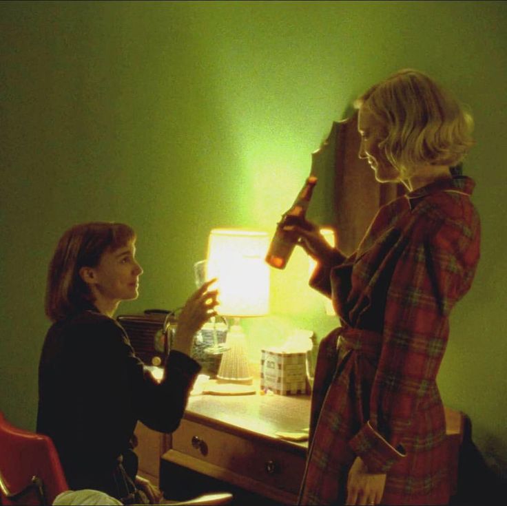 two women standing in front of a table with a lamp on it and one holding a wine bottle