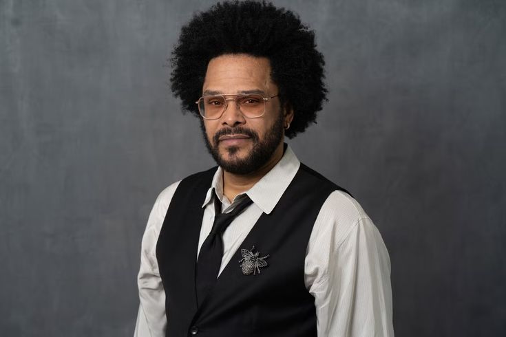 a man with an afro wearing glasses and a black vest is standing in front of a gray background