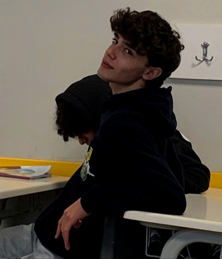 a young man sitting at a desk in front of a computer monitor with another person behind him