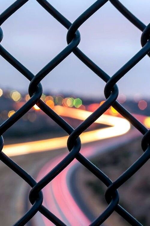 the view from behind a chain link fence looking down at a highway with lights in the distance